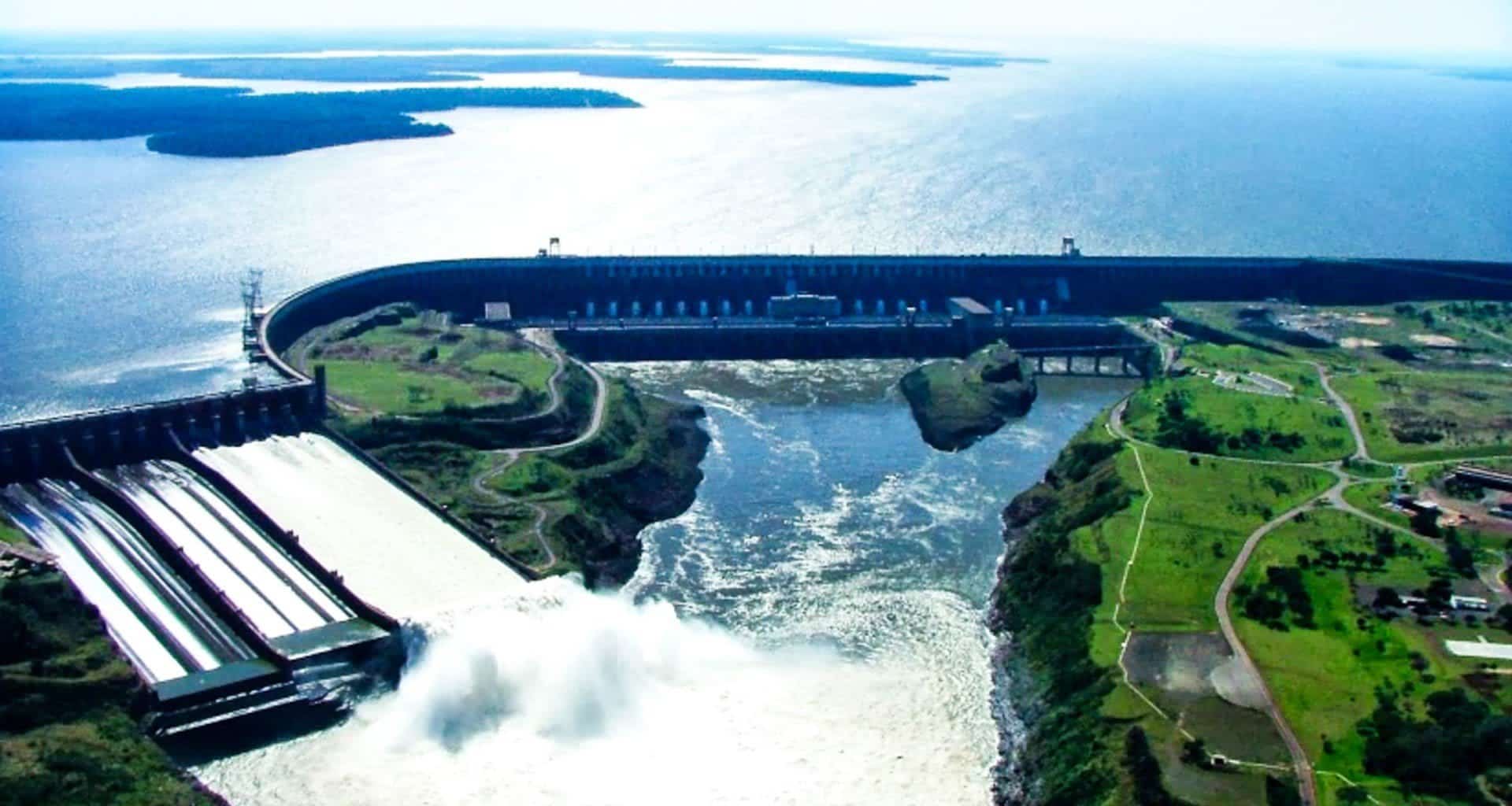 Aerial view of the impressive Itaipu Dam with water flowing through spillways, surrounded by lush green landscape and a vast body of water—an iconic sight on any Paraguay Tour.