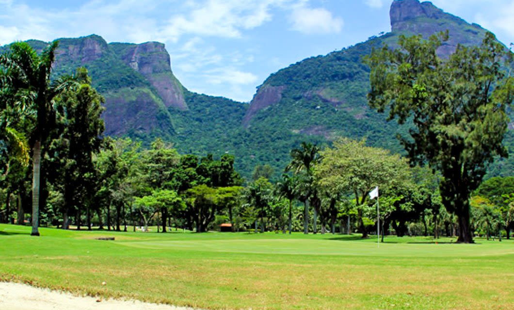 A lush green golf course with a flag on the putting green, surrounded by trees and backed by tall, forested mountains under a blue sky—an ideal setting for an unforgettable Brazil Golf Tour.