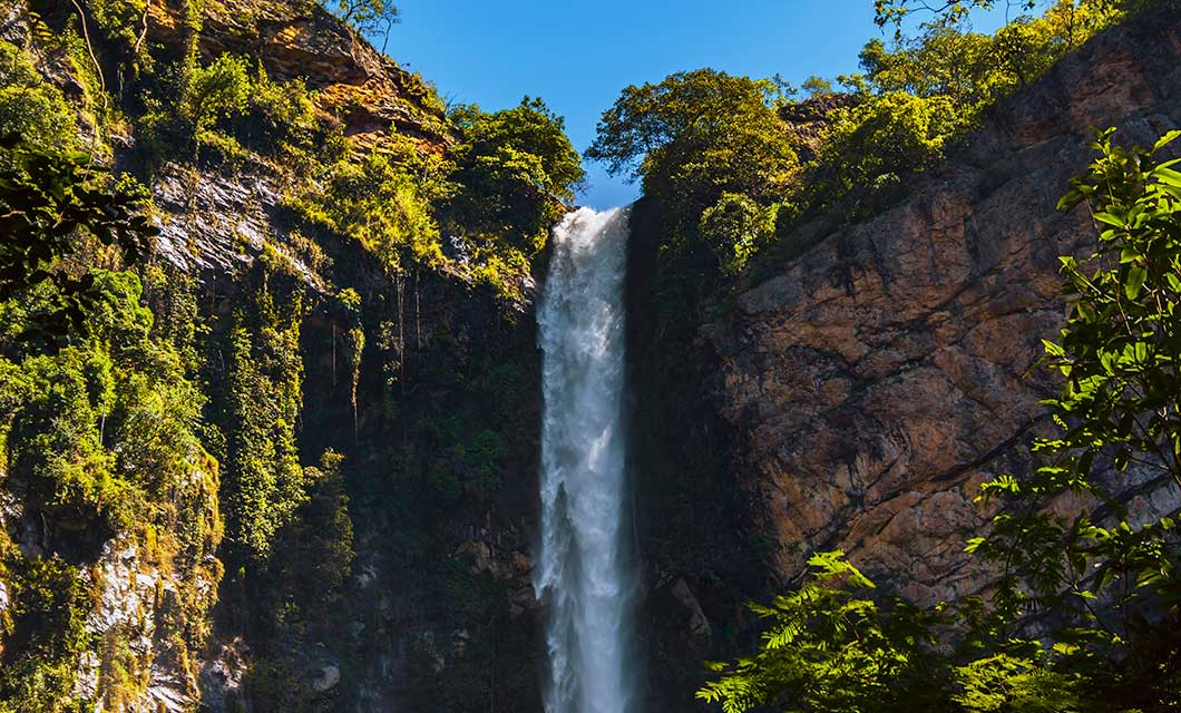 A tall waterfall cascades between rocky cliffs covered in lush green vegetation, surrounded by trees and under a clear blue sky—an iconic scene from Jalapão, deep within Brazil's Cerrado and waiting to be discovered on your next tour.