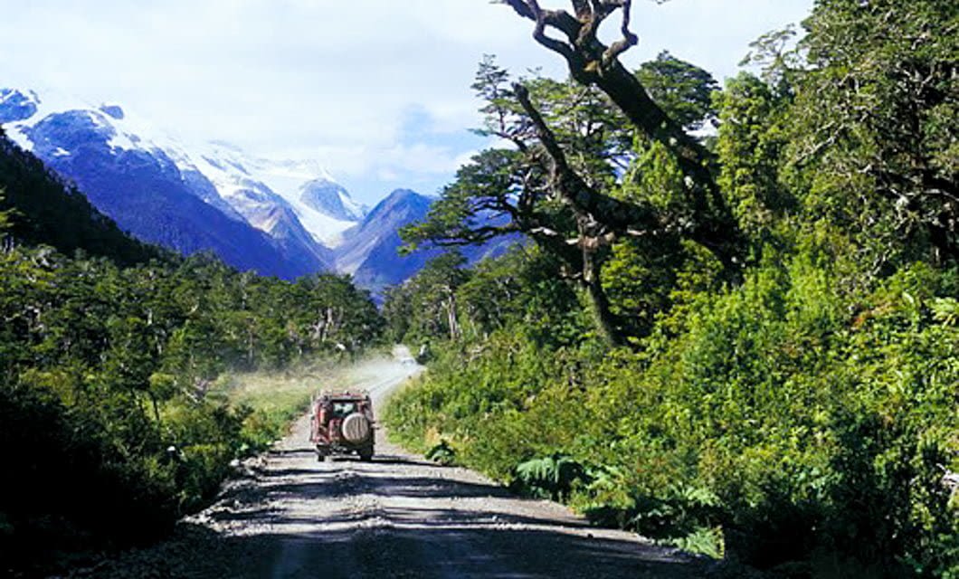 A vehicle drives along a gravel road in Northern Patagonia, surrounded by dense green forest and tall trees, with distant snow-capped mountains under a partly cloudy sky—perfect for an overland adventure.