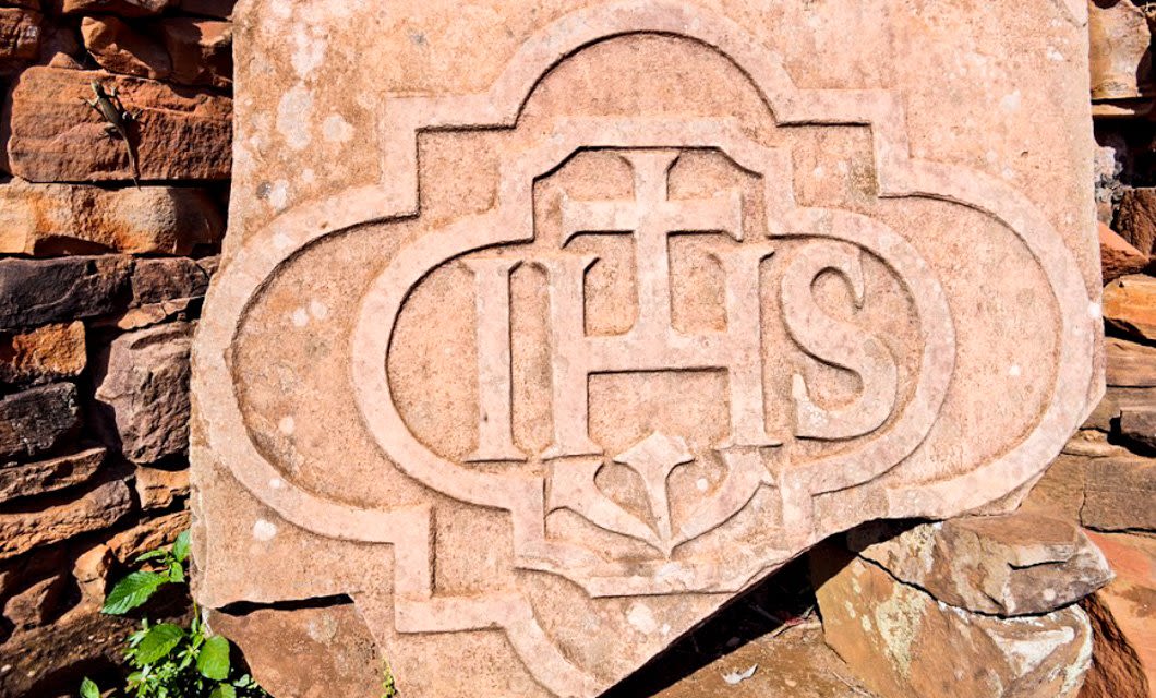 A carved stone slab from the Jesuit Missions features the letters IHS with a cross above the H and a floral design below, set against a decorative border and surrounded by rough stonework.
