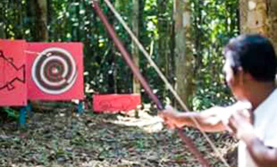 A person aims a bow and arrow at a target with concentric circles, set up on red boards in a forested area reminiscent of the Amazon. The scene is outdoors with trees in the background.