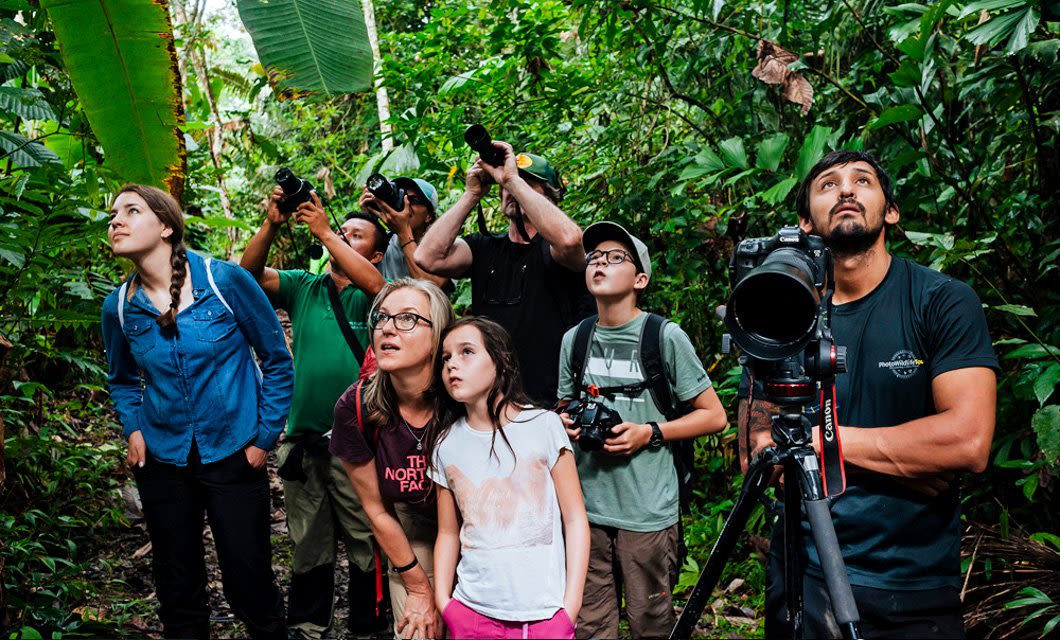 A group of people, including adults and children, stand together in a lush jungle during their Amazon Cruises adventure aboard the Anakonda, looking up with cameras and binoculars as they observe something in the trees amid dense foliage.