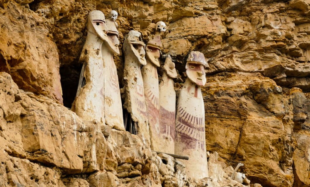 Four large, ancient human-shaped sarcophagi with painted faces and patterns stand against a rocky cliff, evoking the mysterious legacy of the Chachapoyas, with skulls placed above them.