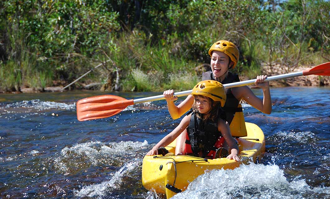 Two people wearing helmets and life jackets paddle a yellow kayak through a shallow, rocky river on a Jalapão tour in Brazil's Cerrado. Both are smiling and appear to be enjoying the adventure surrounded by lush green vegetation.