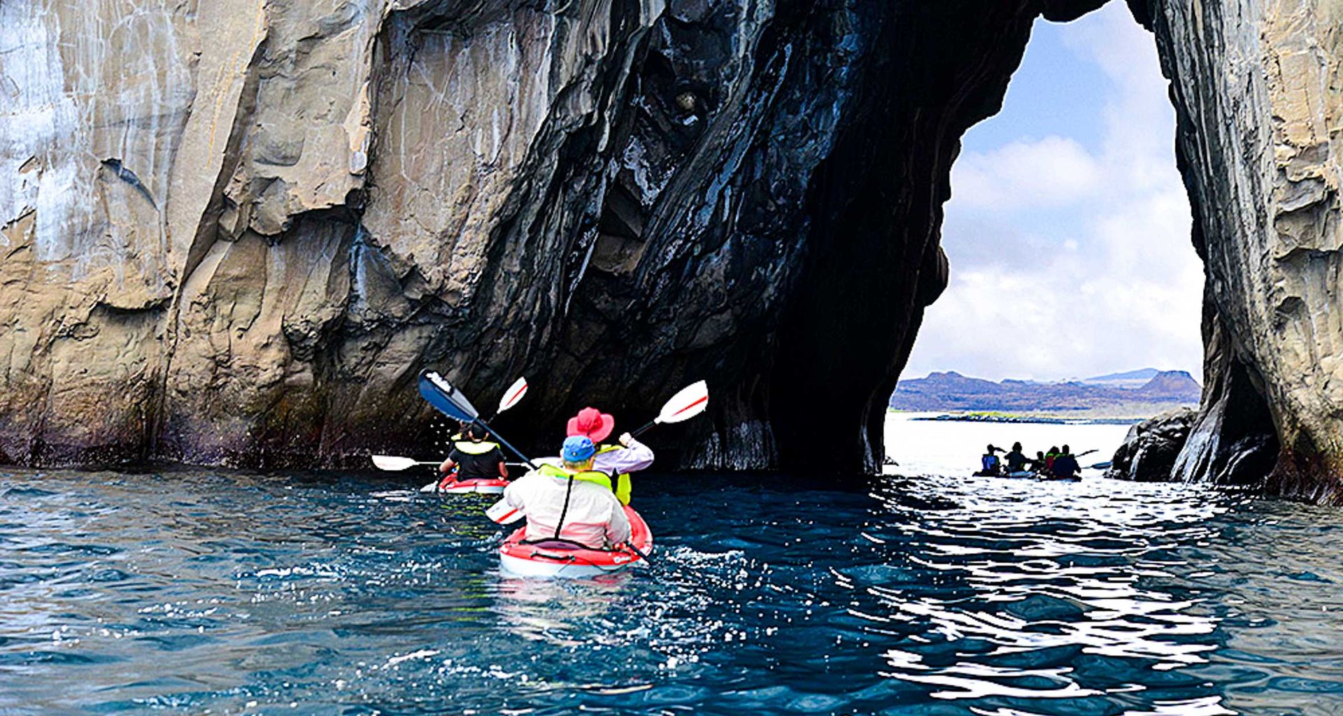 Adventurers on a multisport adventure kayak through a rocky sea arch in the Galapagos, surrounded by blue water and tall cliffs, with distant mountains under a partly cloudy sky.