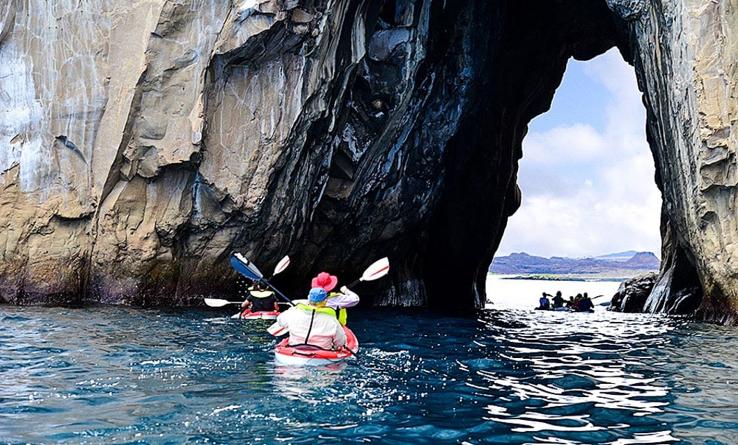 People kayaking through a large rocky sea arch with clear blue water on a Galapagos multisport adventure, heading toward an opening with distant land and a partly cloudy sky visible beyond the arch.