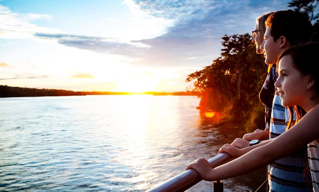 Three people stand by a railing overlooking a wide river at sunset, the sky aglow with warm light—an unforgettable moment aboard the Manatee Explorer as they drift through the heart of the Ecuadorian Rainforest.