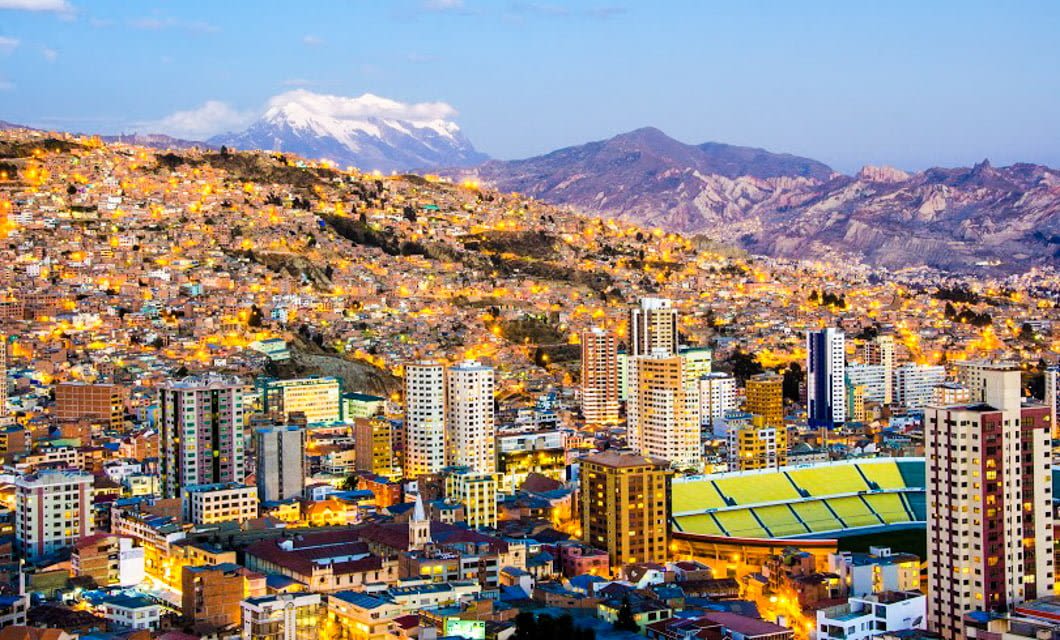 A vibrant cityscape of La Paz, Bolivia at dusk, with many buildings on hilly terrain. City lights glow as a stadium sits in the foreground and snow-capped mountains rise beyond—a perfect view for any La Paz tour under a clear sky.
