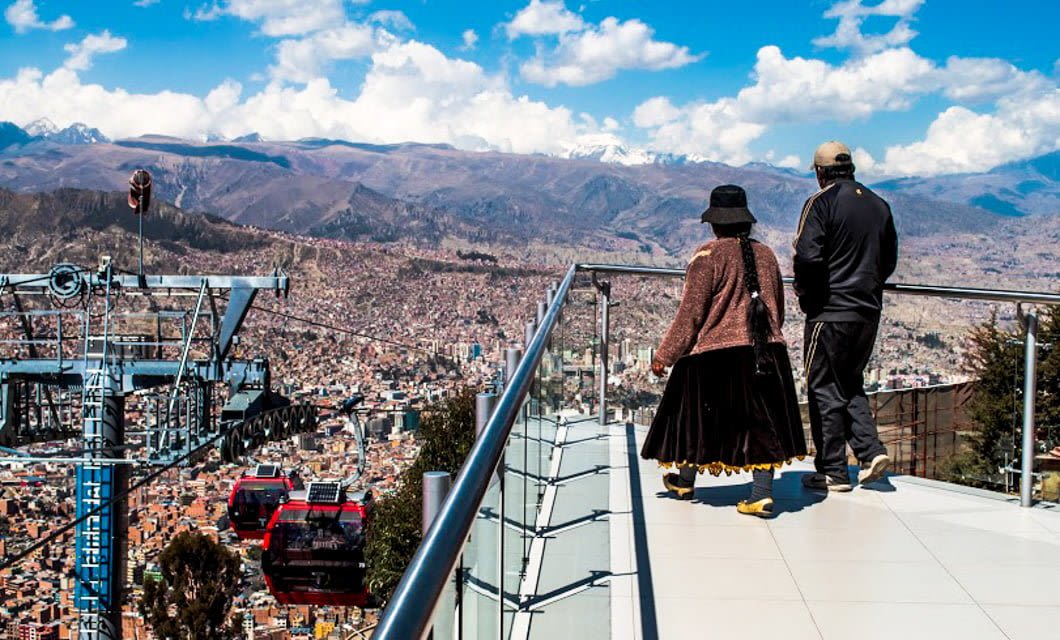 Two people stand on a glass terrace overlooking a sprawling city with mountains in the background—a stunning scene that captures Bolivia highlights, with red cable cars gliding below and a bright sky dotted with scattered clouds.