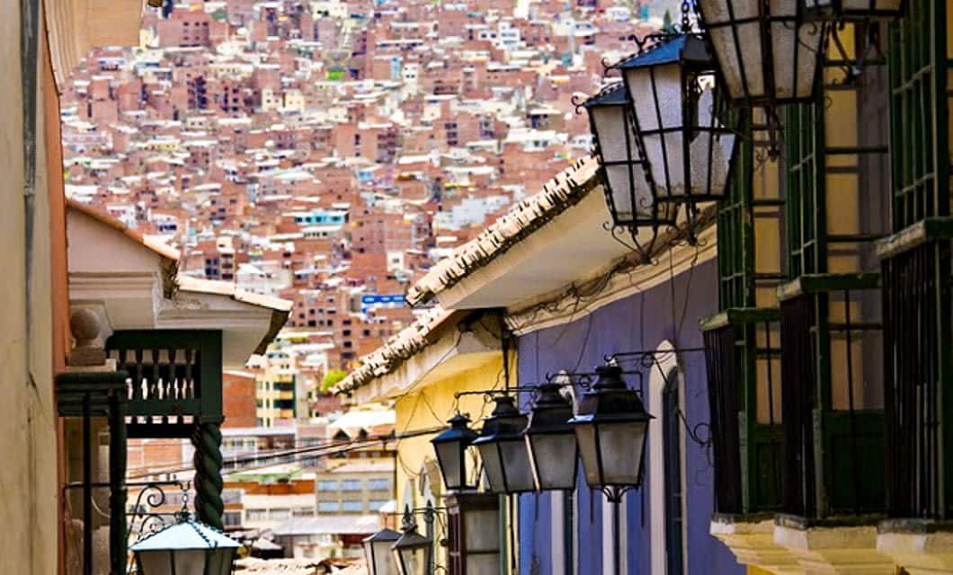 A narrow street in La Paz lined with colorful buildings and hanging lanterns leads to a densely packed hillside filled with many brown, brick houses, echoing the vibrant spirit found along the historic Travels of the Incas.