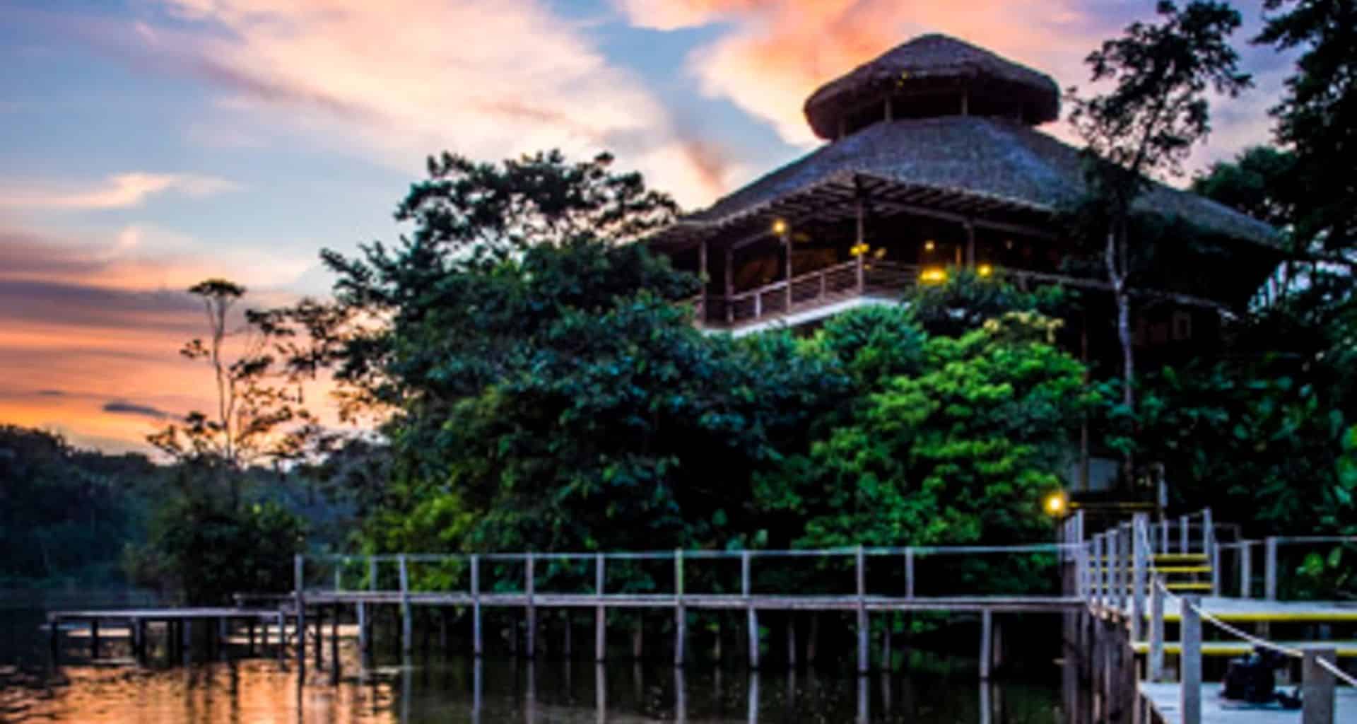 A wooden lodge with a thatched roof—La Selva Amazon Lodge—nestles among lush green trees beside a calm river at sunset, with a wooden walkway leading up to it and colorful clouds painting the Amazon Rainforest sky.