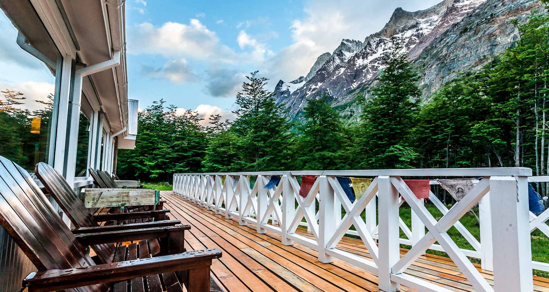 Wooden deck with chairs and a white railing at Lago Grey Lodge overlooks lush green trees and tall, rugged mountains with patches of snow under a partly cloudy sky. The deck is attached to a building with large windows.