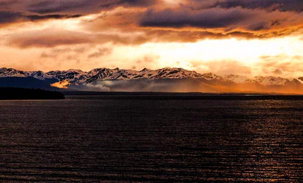 Dramatic sunset over a mountain range with snow-capped peaks, reflected in a dark, rippling body of water—capture the very best of Argentina travel as clouds glow orange and gold across the breathtaking sky.