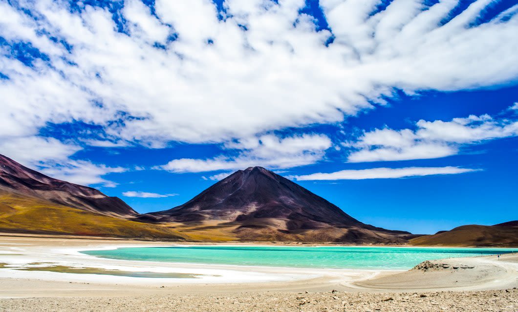 A turquoise lake sits at the base of a dark mountain under a vibrant blue sky with scattered white clouds, surrounded by a dry, sandy landscape near Potosi.