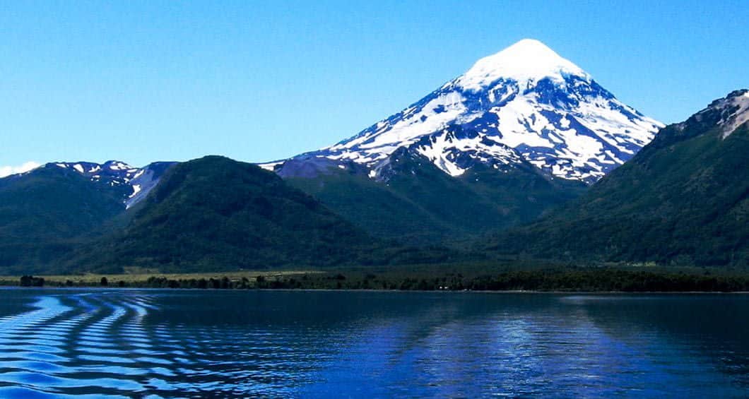 A snow-capped mountain rises above green hills, reflected in the calm, blue water of a lake under a clear sky—a breathtaking view reminiscent of the Argentina to Chile Lake Crossing.