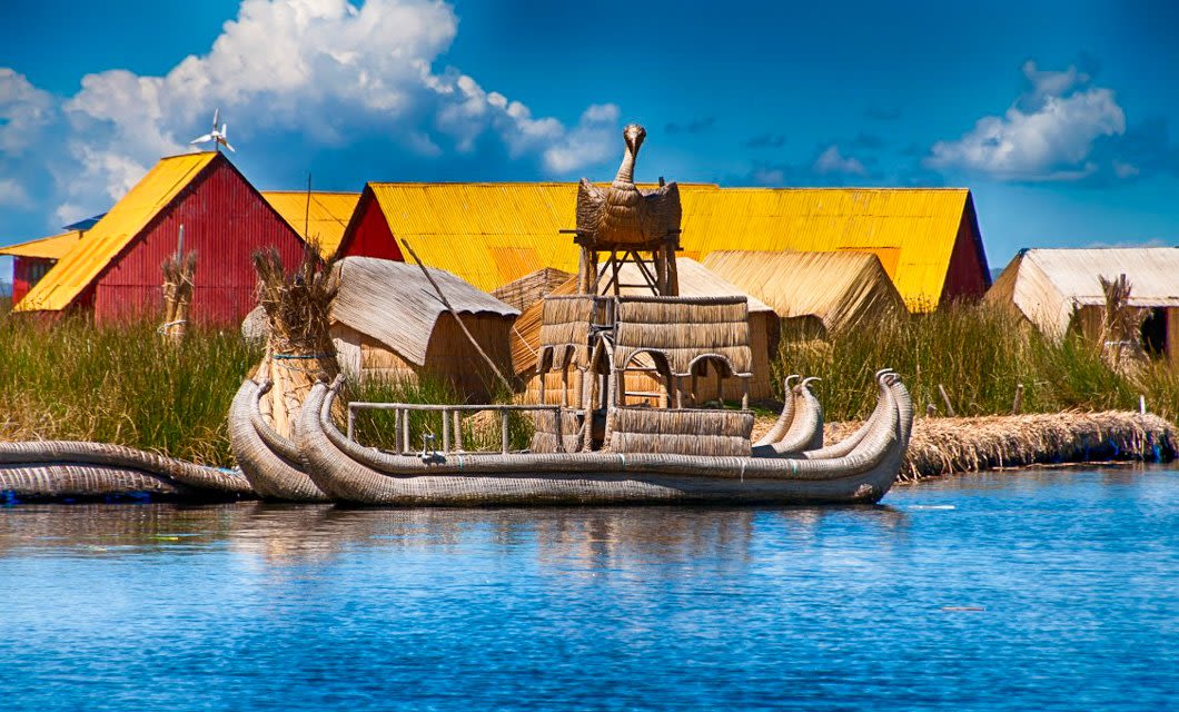 A traditional reed boat floats on clear blue water near straw huts with yellow and red roofs, surrounded by tall grass under a bright, partly cloudy sky—capturing the Essence of Peru and timeless Peruvian culture.