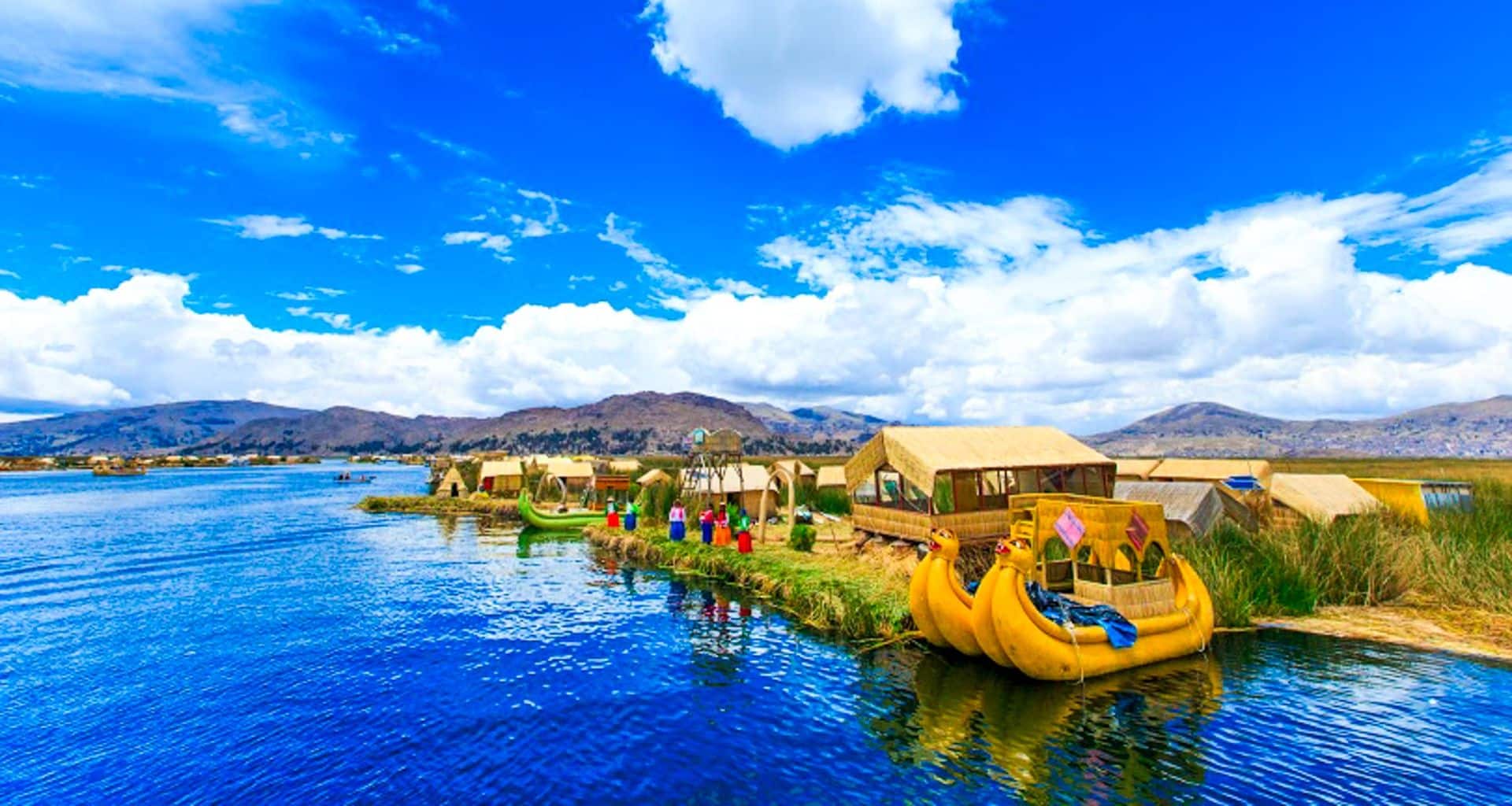 Colorful floating reed huts and boats rest on a vibrant blue lake, with people standing on the reeds—one of the Bolivia highlights you’ll experience during your Bolivia travel adventure, set against mountains and a bright, cloudy sky.