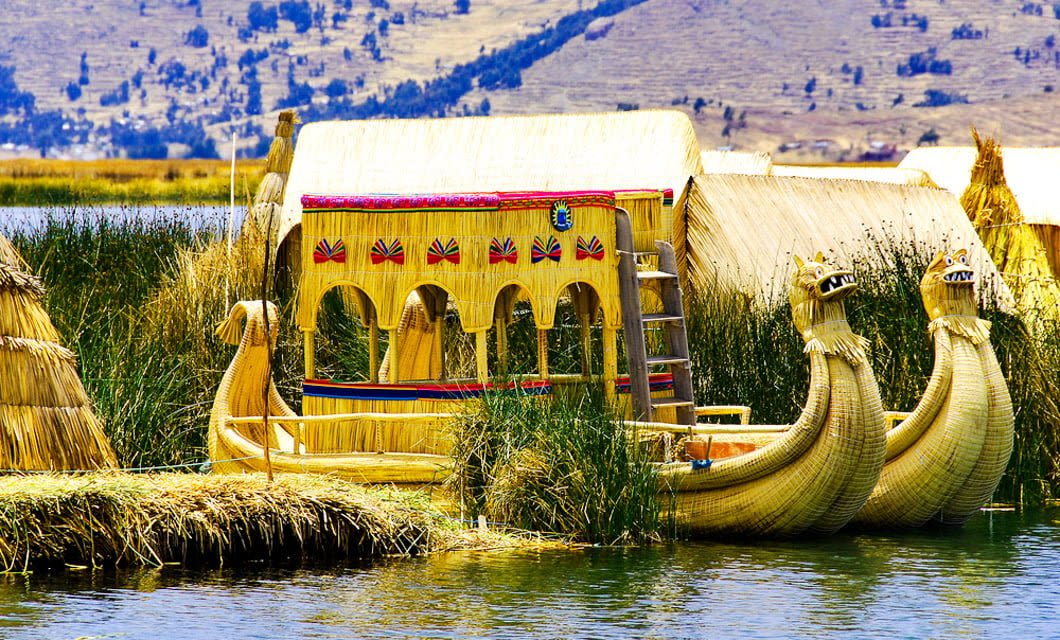 A traditional reed boat with decorative details floats on a lake near reed huts and tall grasses, with mountains visible in the background—a scene reminiscent of the ancient Travels of the Incas.