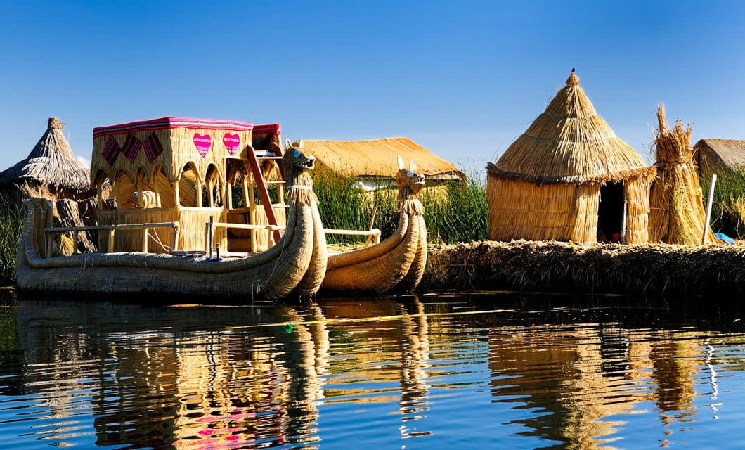 Traditional reed boat with ornamental arches and thatched reed huts on the water’s edge, reflected in calm water under a clear blue sky. Lush green reeds grow behind the huts—a peaceful scene to enjoy before your Peru golf tour adventure.
