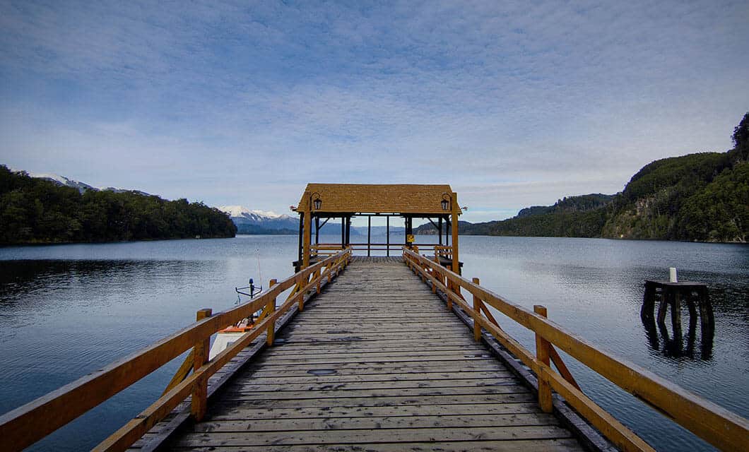 A wooden pier with railings extends over a calm lake toward a covered shelter, surrounded by forested hills and distant snowy mountains under a partly cloudy sky, capturing the tranquil beauty of Wild Argentina.