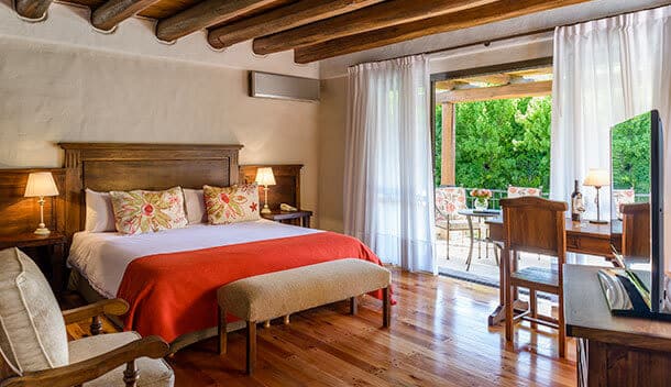 A cozy bedroom with a wooden bed, floral pillows, orange throw, armchair, desk, and TV. Sunlight streams through sheer curtains, opening to a patio with chairs and lush greenery—the perfect honeymoon retreat in the Sacred Valley.