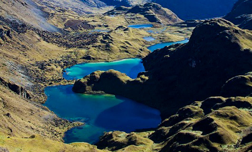 A highland landscape showing a series of vibrant blue lakes surrounded by rugged, grassy mountains and rocky terrain under daylight, capturing the stunning scenery along the Lares Valley Trek in Peru.