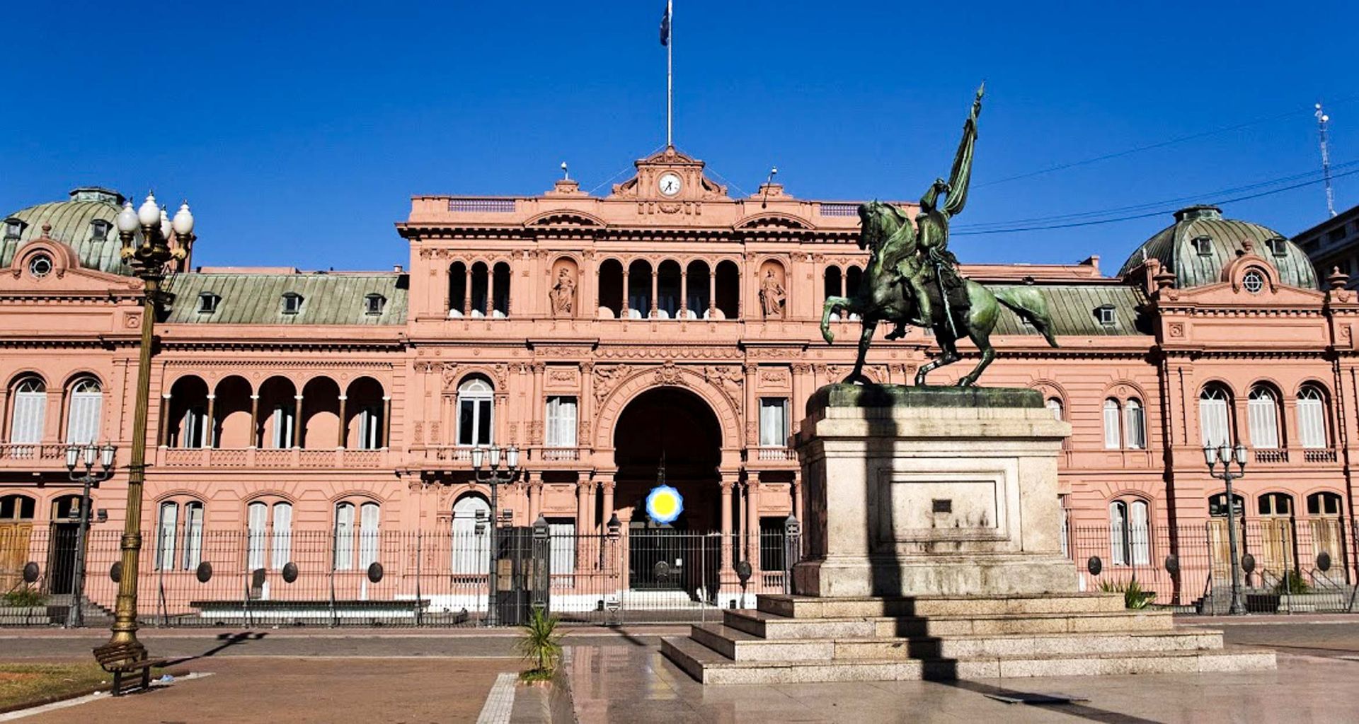 A grand pink government building in Buenos Aires with arched windows and ornate details, featuring a statue of a rider on horseback in the foreground, perfect to admire during a city tour under the clear blue sky.