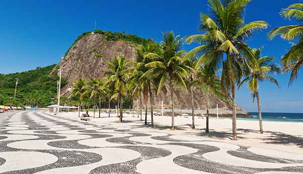 A beachside walkway lined with palm trees and a wavy black-and-white tile pattern stretches along beaches of golden sand, with a large rocky hill in the background under a clear blue sky.