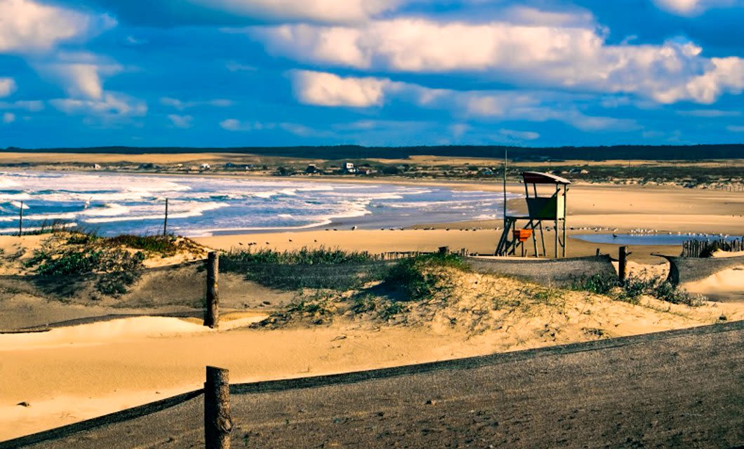 A sandy beach in Uruguay with gentle waves, grassy dunes, a wooden lifeguard tower, and distant houses under a bright blue sky—perfect for leisurely walks or even horseback riding along the shore.