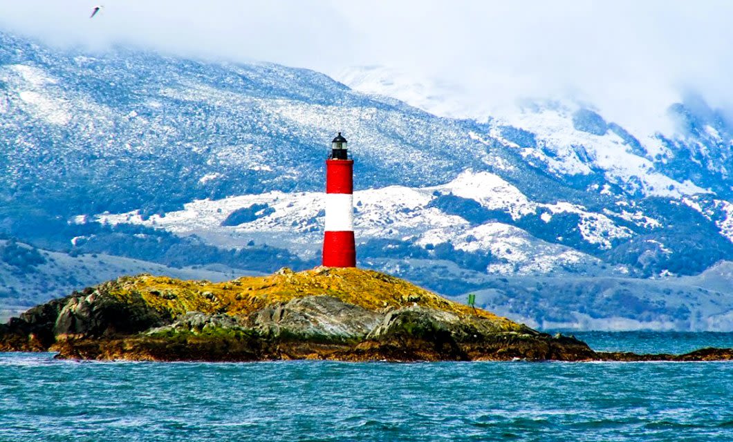 A red and white striped lighthouse stands on a rocky island surrounded by water, snow-capped mountains in the background—an iconic sight on a Buenos Aires to Tierra del Fuego cruise under a dramatic cloudy sky.