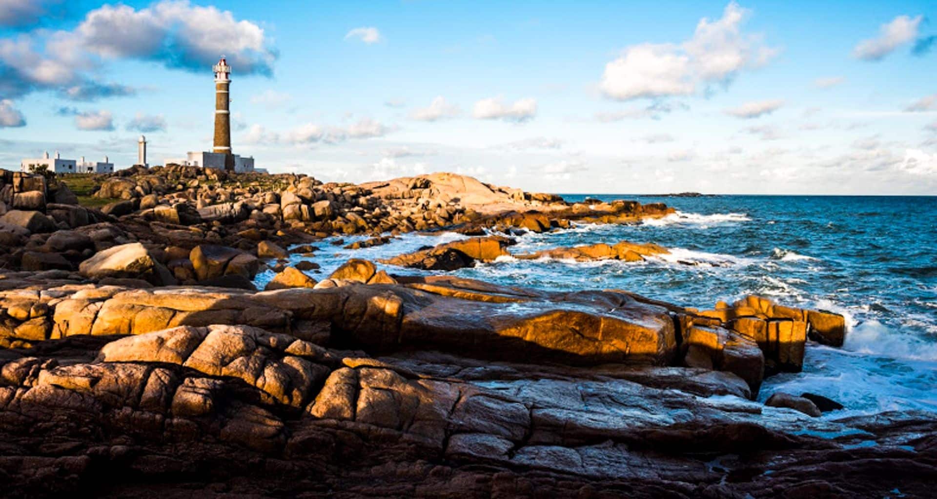 A tall lighthouse stands on Uruguay’s rocky coastline under a blue sky with scattered clouds, with waves crashing against the rocks and the ocean stretching out to the right—perfect scenery for an unforgettable horseback riding adventure.