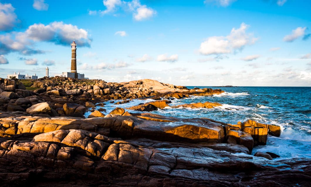 A tall lighthouse stands on rocky cliffs overlooking the ocean in Uruguay, with waves crashing below and sunlight casting warm tones—an ideal backdrop for a scenic horseback riding adventure along the coast.