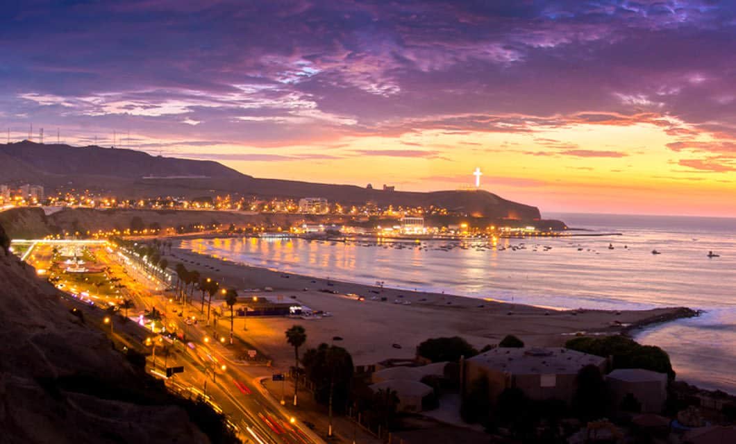 A coastal city at sunset with a curving shoreline, bright city lights, a sandy beach, and a large illuminated cross on a distant hill under a colorful Lima sky.