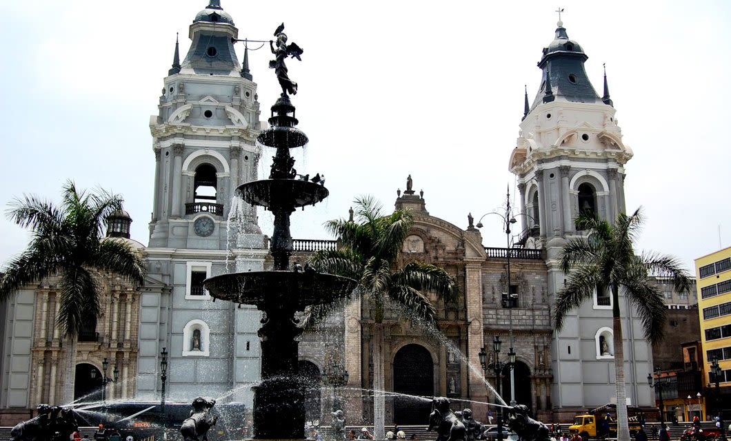 A historic plaza in Lima with a large tiered black fountain in the foreground, framed by palm trees, and a grand cathedral with two tall towers in the background under a cloudy sky.