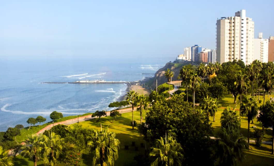 A coastal cityscape featuring tall buildings, lush green park areas with palm trees, and a winding path overlooking the ocean under a clear blue sky—inviting as an Amazon Lodge after a Machu Picchu Tour.