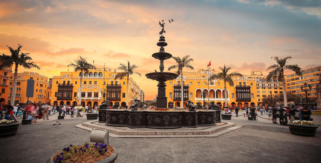 A historic fountain surrounded by people stands in the center of a plaza with yellow colonial-style buildings and palm trees at sunset, capturing the vibrant charm of one of South America's cultural capitals under a colorful orange and blue sky.