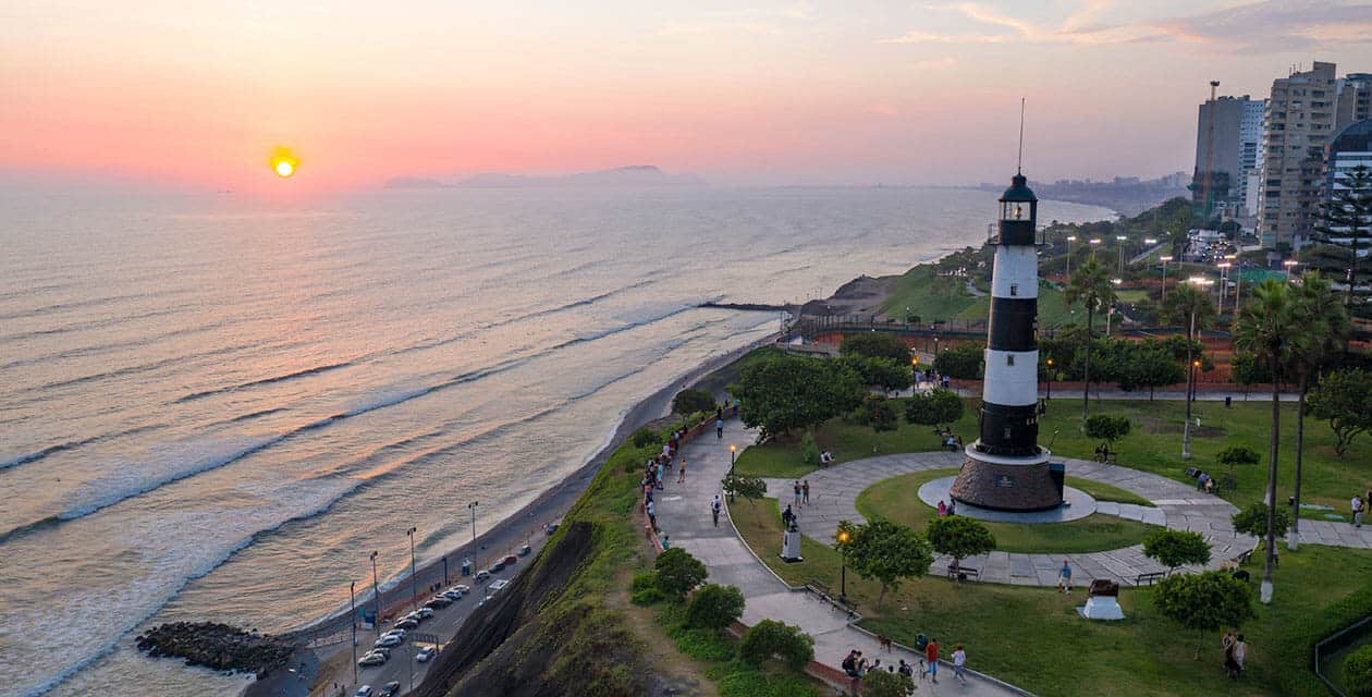 A black and white lighthouse stands on a green park atop cliffs overlooking the ocean at sunset, with people walking nearby and city buildings in the background—a scenic gem among South America's natural wonders, bathed in the sun's warm glow.