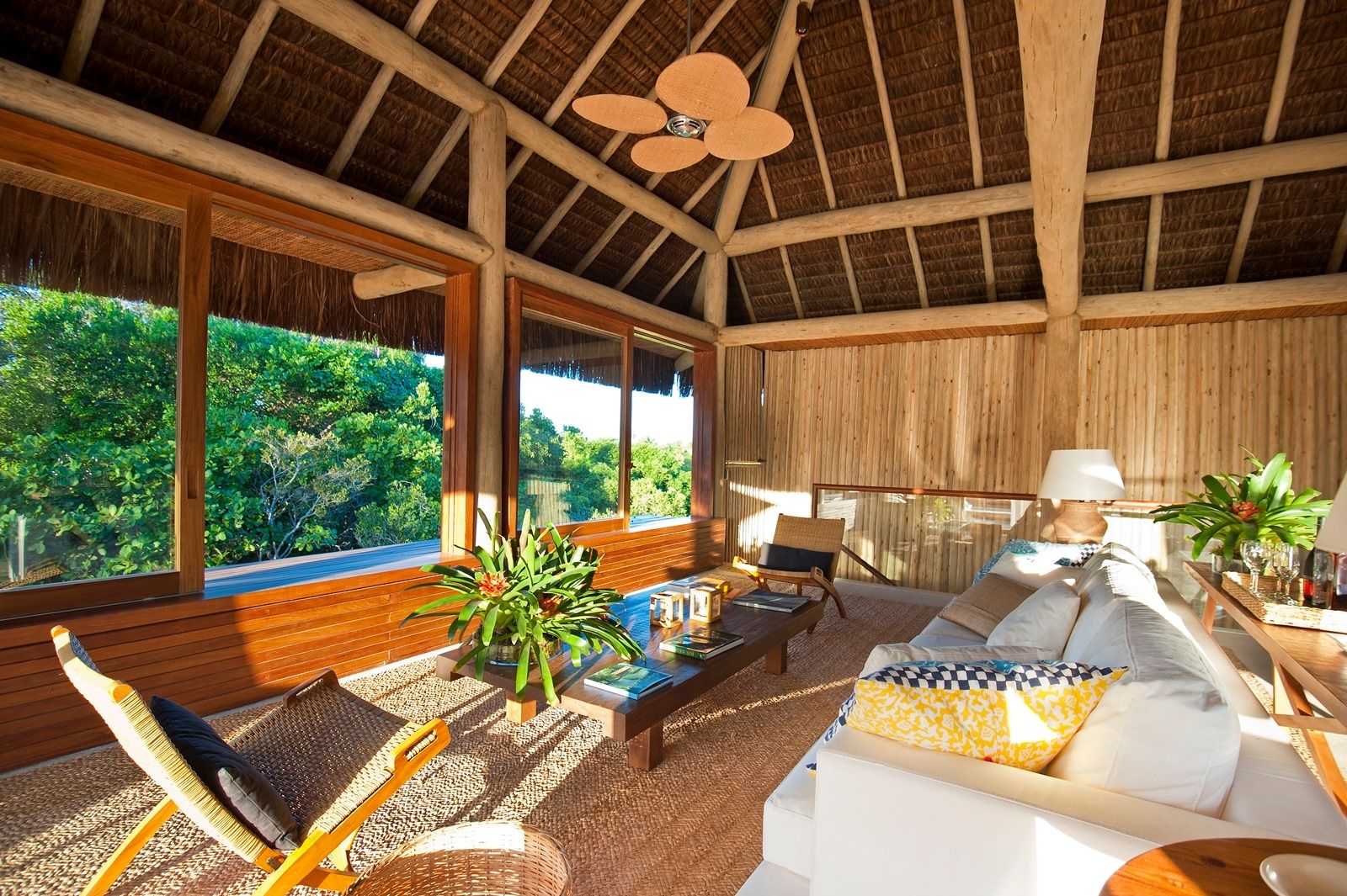 A bright, tropical living room inspired by Praia do Forte’s eco-resort style, with wooden walls and ceiling, large windows showcasing greenery, wicker furniture, a white sofa with yellow pillows, indoor plants, and sunlight streaming in.
