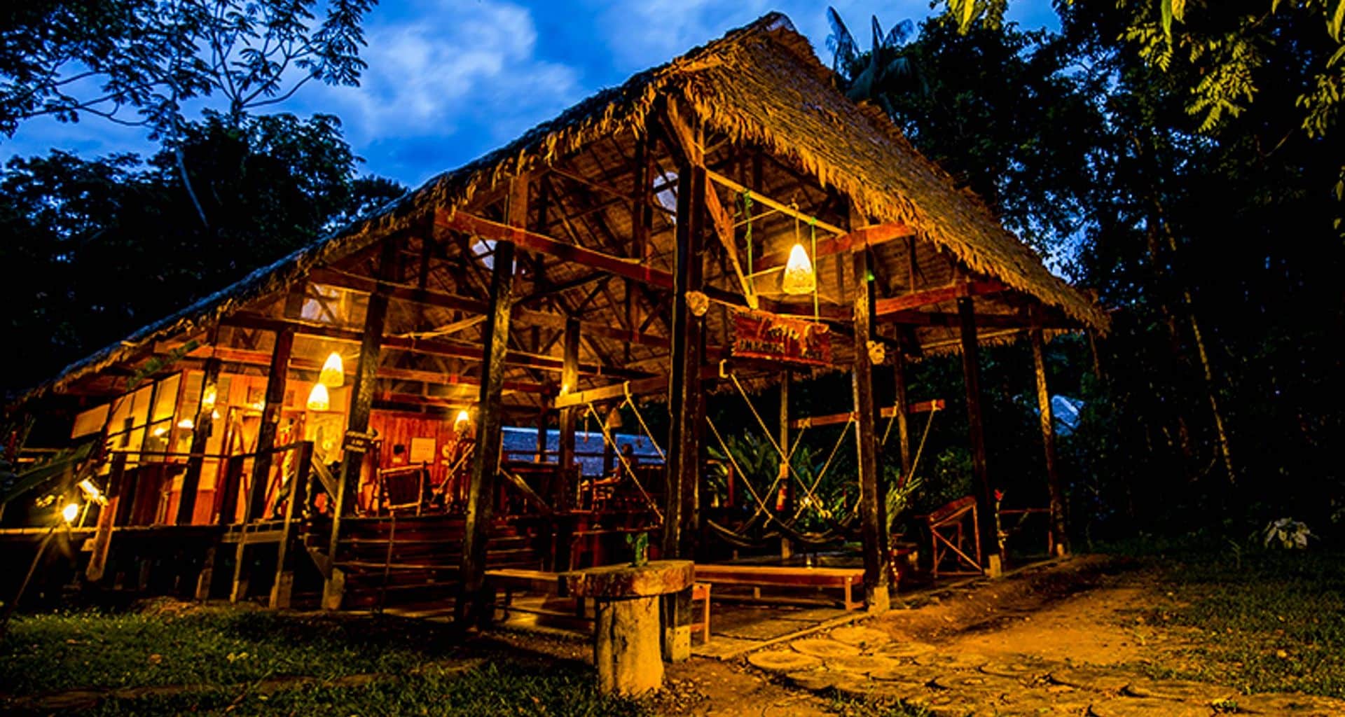 A rustic, open-air Amazon Lodge with a thatched roof, warmly lit by hanging lamps, sits surrounded by lush trees under a dusky, cloud-streaked sky—an inviting retreat in the heart of the Peru Amazon Lodge experience.