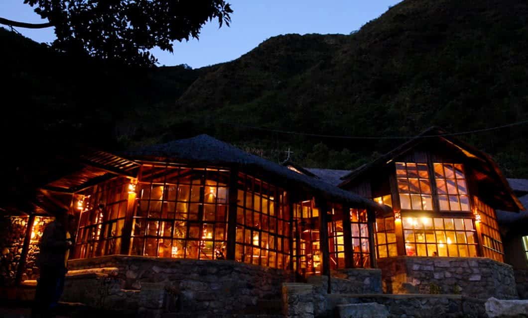 A warmly lit wooden and glass Salkantay Lodge with stone walls sits at the base of a mountain at dusk, glowing through large windows against the darkening sky and surrounding trees—a serene stop on your Lodge to Lodge Trek.