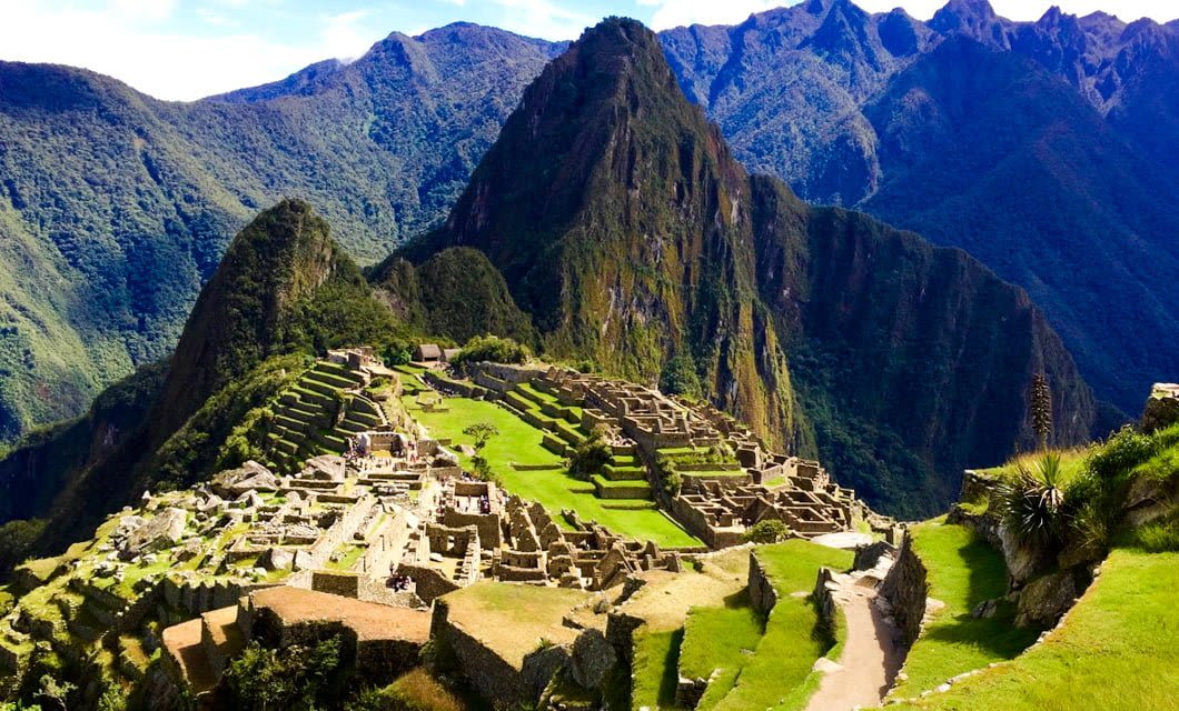 Ancient stone ruins of Machu Picchu sit atop a lush, green mountain ridge, surrounded by steep, forested peaks under a blue sky—a must-see on any South America tour.