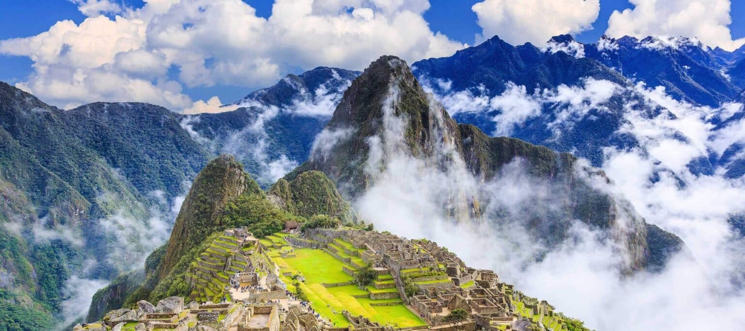 Ancient stone ruins of Machu Picchu sit atop a lush mountain ridge, surrounded by green peaks and drifting clouds, making it a breathtaking highlight of many Peru tours under the vivid blue sky.