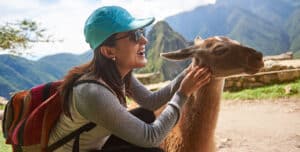 A smiling woman wearing sunglasses, a blue cap, and a colorful backpack pets a llama outdoors with mountains and ruins in the background, capturing the spirit of a Perfect Trip during her Peru travel adventure.