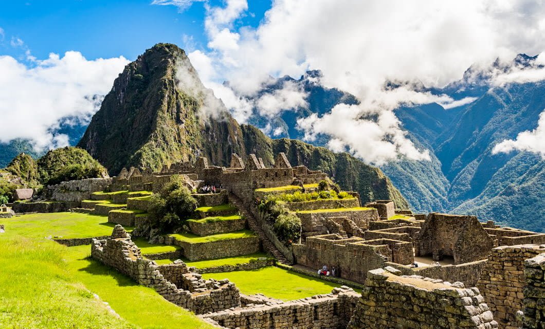 Ancient stone ruins of Machu Picchu, the essence of Peru, sit atop lush green terraces, surrounded by mountains and clouds under a bright blue sky.