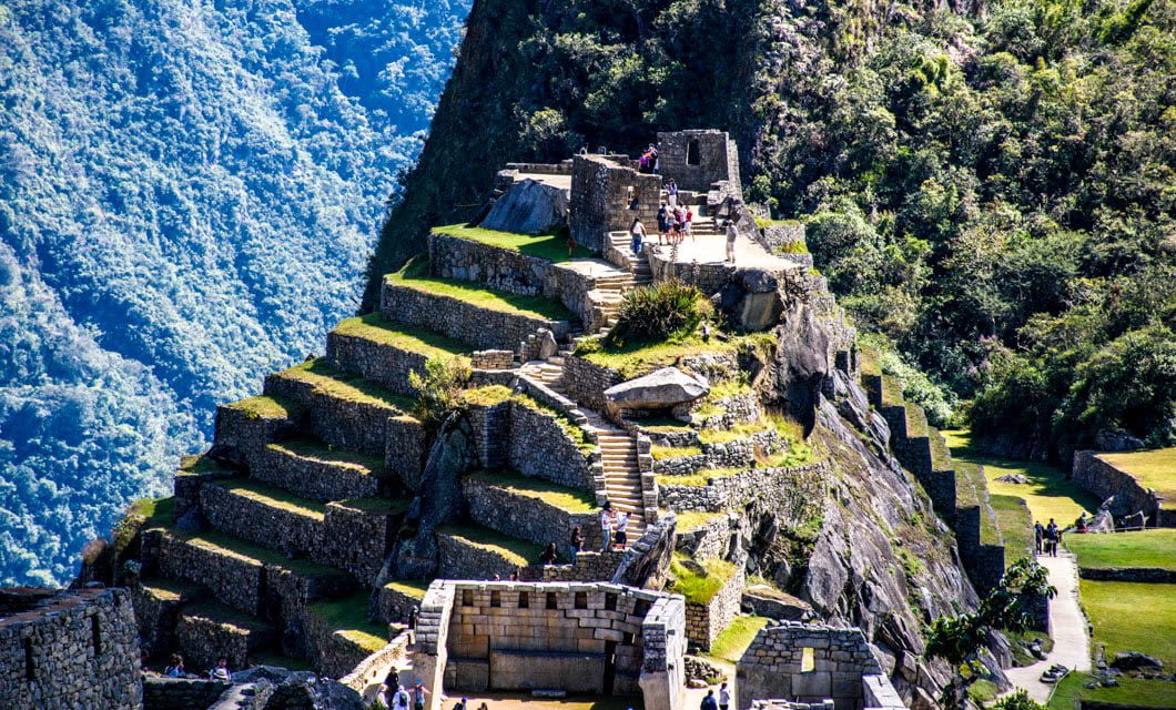 Ancient stone ruins of Machu Picchu are set on terraced green hillsides with mountains in the background. Several tourists explore the historic site under bright sunlight on a memorable South America Tour.
