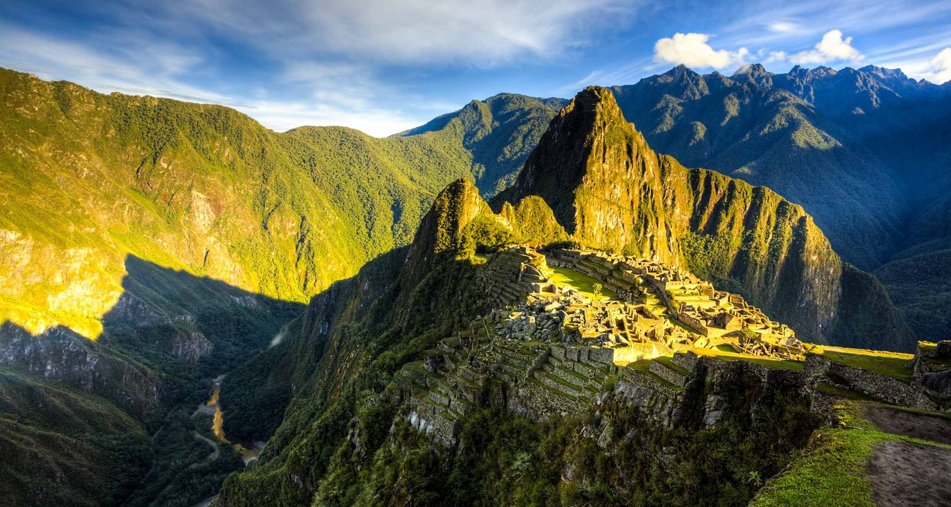 Ancient stone ruins of Machu Picchu sit atop a lush, green mountain ridge near Cuzco, under a bright, partly cloudy sky, with dramatic sunlight and deep shadows highlighting the surrounding peaks.