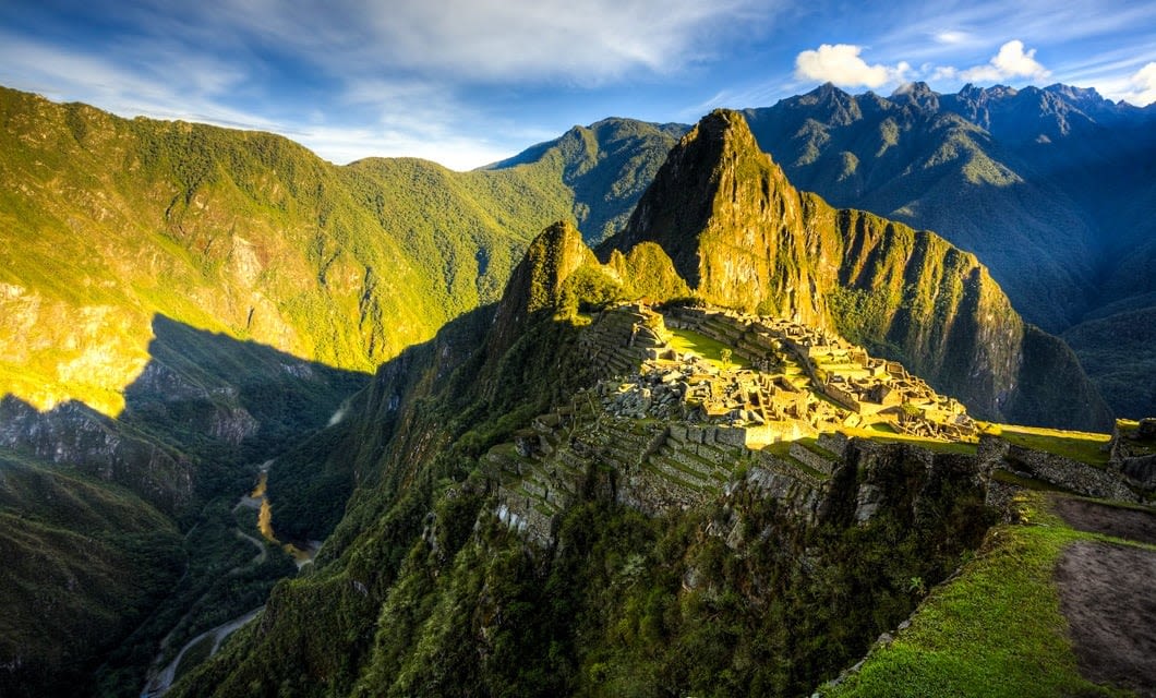 Ancient stone ruins of Machu Picchu sit atop a lush green mountain ridge in Peru, where the legendary Inca Trail ends, surrounded by dramatic peaks under a bright, partly cloudy sky. Sunlight highlights part of the archaeological site.