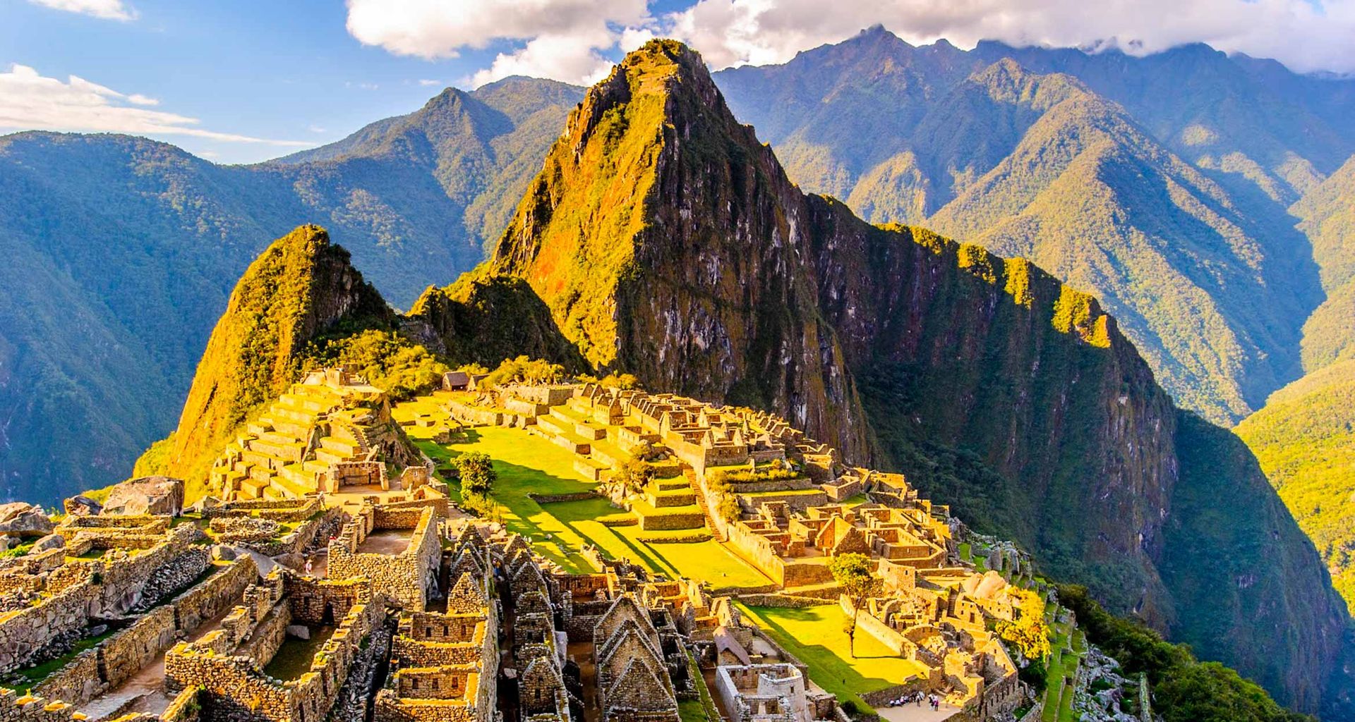Ancient stone ruins of Machu Picchu sit atop a green mountain ridge, surrounded by lush, steep peaks under a partly cloudy sky in the Andes of Peru, often explored on a Lodge to Lodge Trek like the renowned Salkantay Trek.