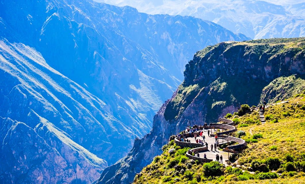 A winding paved path with tourists captures the essence of Peru as it overlooks a deep, green canyon with steep, rugged mountain walls under bright sunlight.